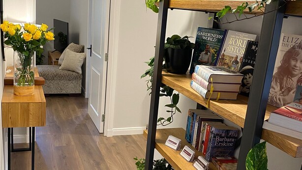 A hallway with a wooden floor, a bookshelf filled with books, a potted plant, and a vase with yellow flowers. - TravelNess Apartment (Inverness)