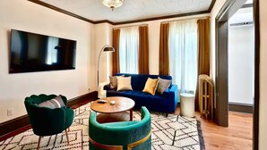 A modern living room with a blue sofa, green armchairs, a round coffee table, and a flat-screen TV mounted on the wall.