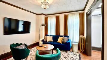 Un salon moderne avec un canapé bleu, des fauteuils verts, une table basse ronde et un téléviseur à écran plat fixé au mur.