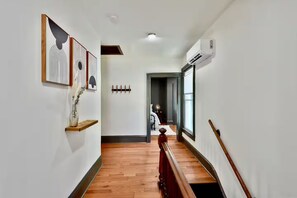 A hallway with wooden flooring, a staircase with a wooden handrail, and a wall-mounted air conditioning unit.