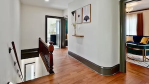 A hallway with wooden flooring, a staircase with a wooden handrail, and a door leading to a room with a blue sofa and a patterned rug.