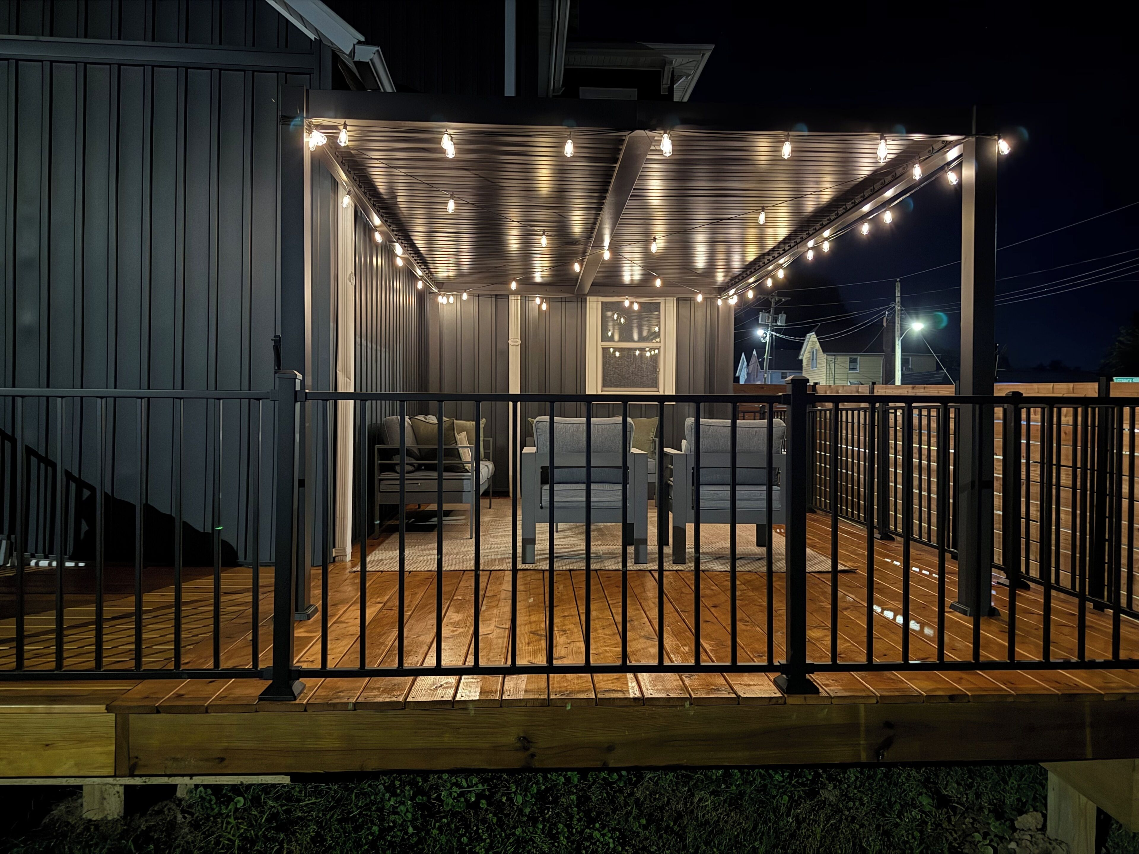 Un espace extérieur abrité, doté d’une terrasse en bois, d’une rambarde en métal et illuminé par des guirlandes lumineuses.
