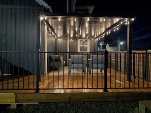 Un espacio de estar al aire libre techado con luces colgantes, una terraza de madera y una barandilla de metal.