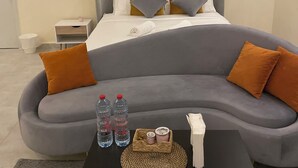 Et moderne hotelværelse med en buet grå sofa, to orange puder, et lille sengebord med en hvid lampe og et sort sofabord med to vandflasker og en kurv med snacks.