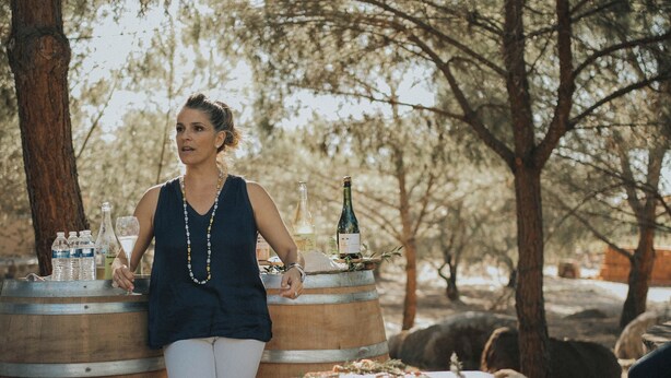 Une femme se tient près d’une table, à côté de laquelle se trouvent des barils de nourriture et de vin, dans un espace boisé.