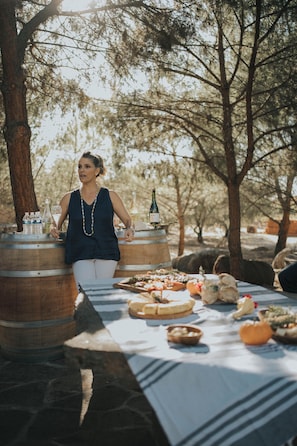 A woman standing by a table with food and wine barrels in a wooded area. - La Mata de Nuez (Valle de Guadalupe)