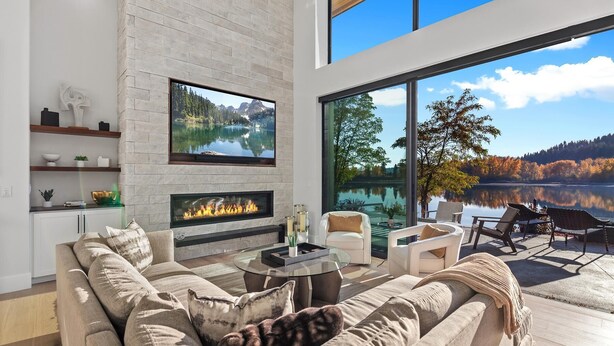 A modern living room with a fireplace, a large TV, and a view of a lake and forest.