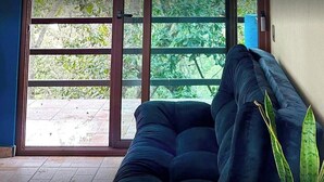 A room with a large sliding glass door leading to a patio. There is a dark blue sofa and a potted plant on the right side. The text 'Rancho Paraíso Real' is visible on the wall.
