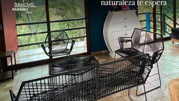 Interior moderno com mobiliário de metal preto, grandes janelas com vista para vegetação, e informações de contato para Rancho Paraíso Real.