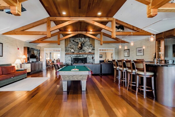 A spacious living area with a pool table, wooden ceiling beams, a stone fireplace, and a bar area with stools.