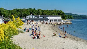 A beach with people relaxing and playing, a large building with a dock, and a forested hill in the background. - Resort style, family friendly getaway! (Lincolnville)
