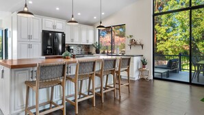 A modern kitchen with white cabinets, a black refrigerator, wooden bar stools, pendant lights, and a large window overlooking a deck. - Luxury Desert Retreat w/ Hot tub + Fire Pit! (Sedona)