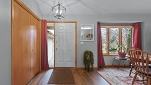 Un pasillo con puerta de madera, alfombra, una silla, una planta en maceta y ventana con cortinas.