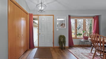 Un couloir avec une porte en bois, un tapis, une chaise, une plante en pot et une fenêtre avec des rideaux.