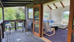 A wooden deck with outdoor seating, a sliding glass door leading to a living area with a sofa and a coffee table. - Charming Beach House (Ruby Bay)