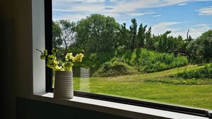 A window with a view of a grassy area and trees, with a white vase with flowers on the window sill. - Charming Beach House (Ruby Bay)