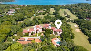 An aerial view of a resort with multiple buildings, a golf course, and a large body of water in the background. - Treetop Ocean View Penthouse - Jobo 15 (Brasilito)