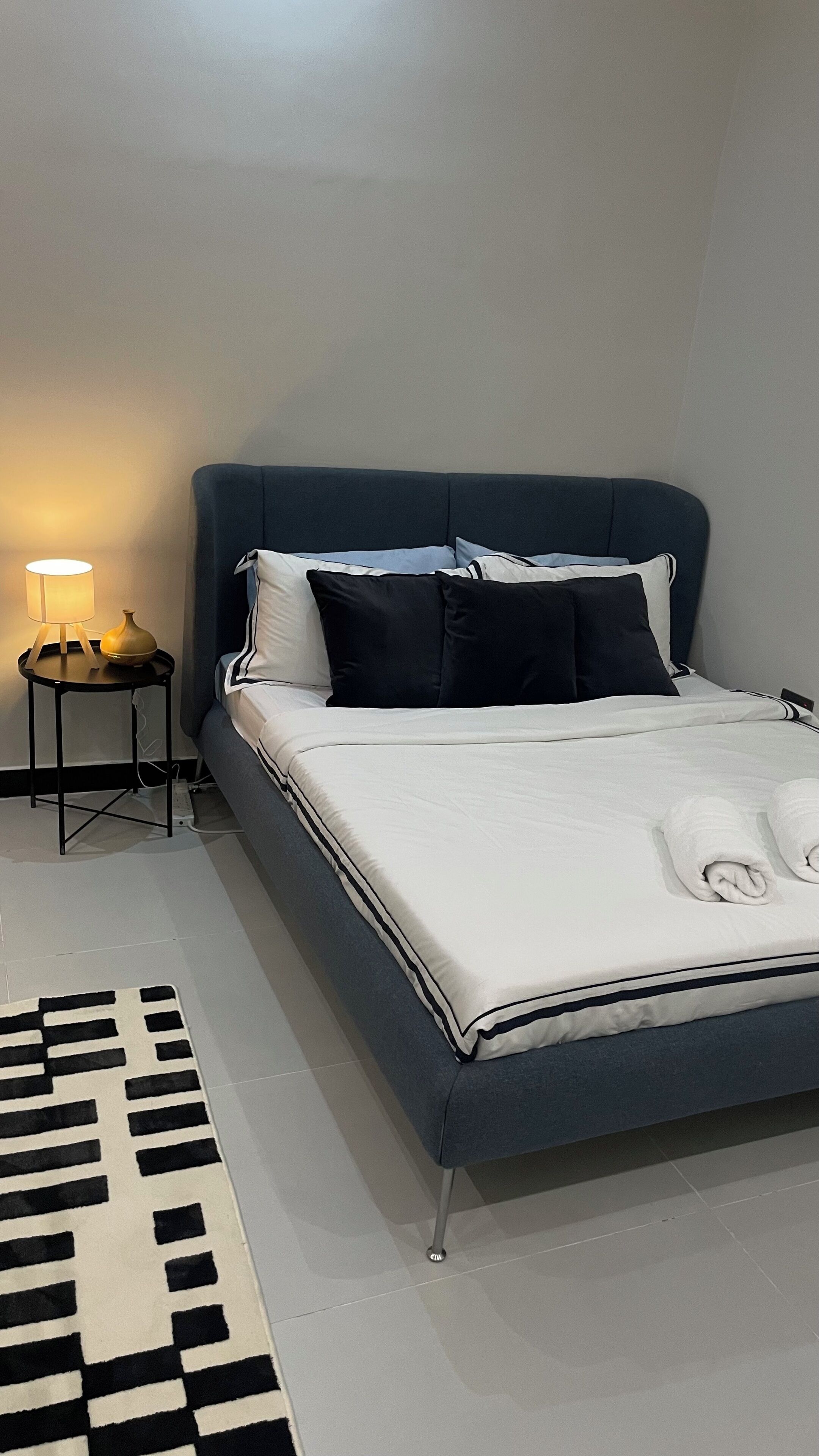 Una cama bien tendida con un cabecero oscuro, una lámpara de noche y una alfombra blanco y negro sobre suelo de baldosas.