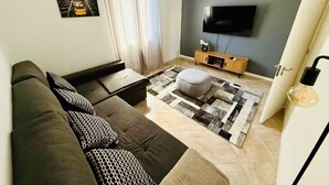 Uma sala de estar moderna com um sofá escuro, um tapete estampado, um pequeno banquinho, um rack de TV de madeira e uma televisão de tela plana grande fixada na parede.