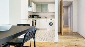 Une cuisine moderne avec des meubles blancs, un four à micro-ondes, un four et un lave-linge. On y trouve une table à manger avec des chaises et un tapis à motifs.