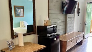 Ett rum med ett skrivbord i trä, ett svart kylskåp, en spegel, en tv som är monterad på väggen och en säng med vita sängkläder.