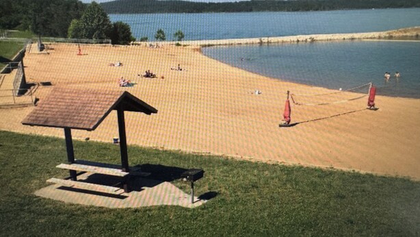 Ein Strand mit Picknicktisch und Sonnenschirmen, ein See im Hintergrund und eine Wiese im Vordergrund.