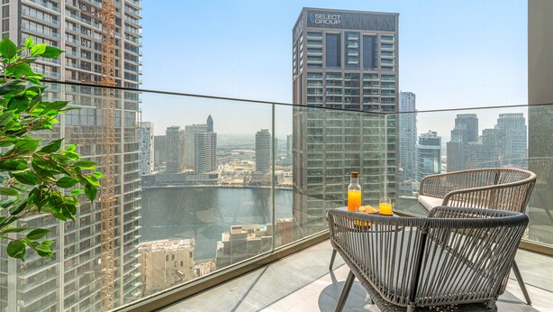 Een balkon met uitzicht op een stadsbeeld en een rivier. Er staan twee rieten stoelen en een tafel met een fles en een glas.