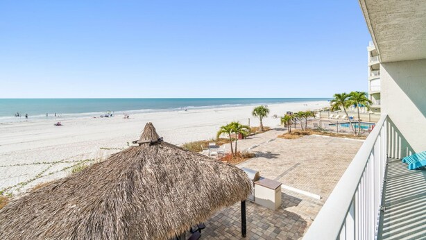 Une vue sur la plage depuis un balcon agrémenté d’un parasol en chaume, de palmiers et d’un espace piscine.