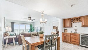 Une salle à manger spacieuse, avec une table et des chaises en bois, un ventilateur de plafond et une vue sur l’océan grâce à de grandes fenêtres.