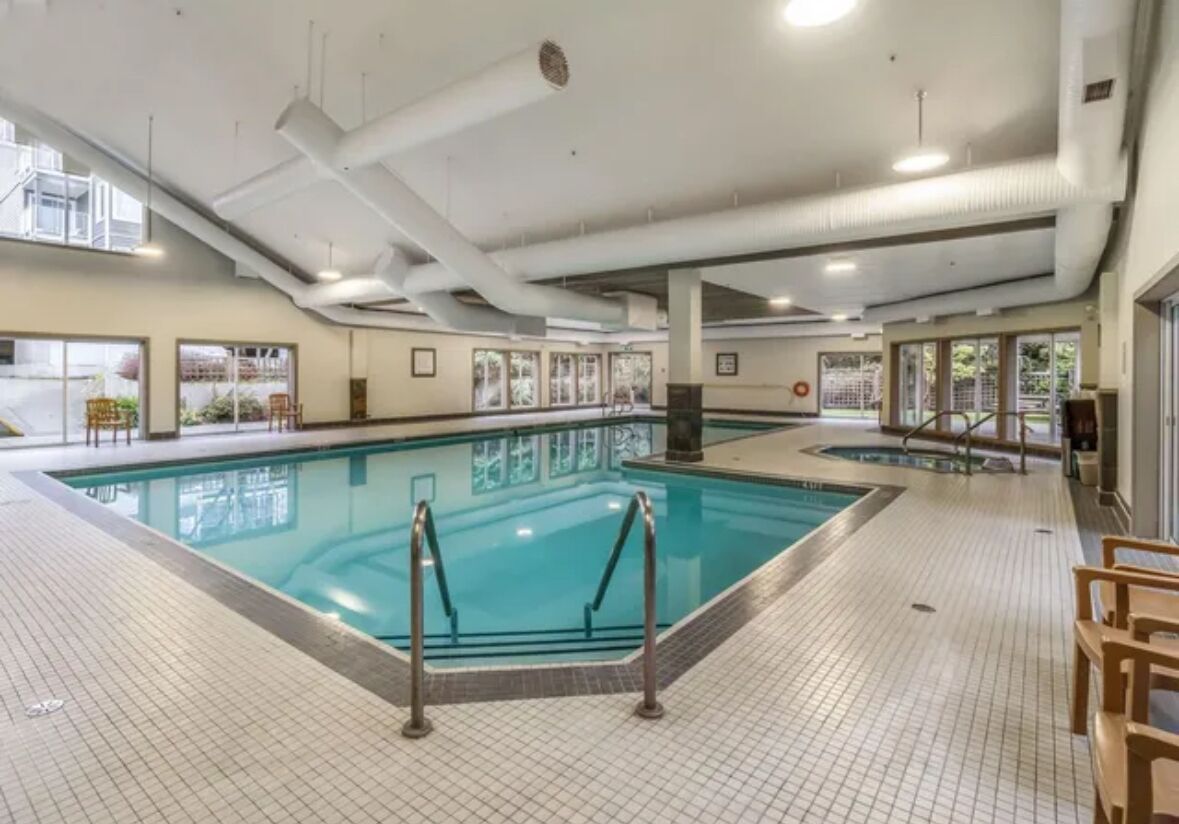 Une piscine intérieure dotée d’un sol carrelé, de rampes en verre transparent et éclairée par la lumière naturelle provenant de fenêtres.