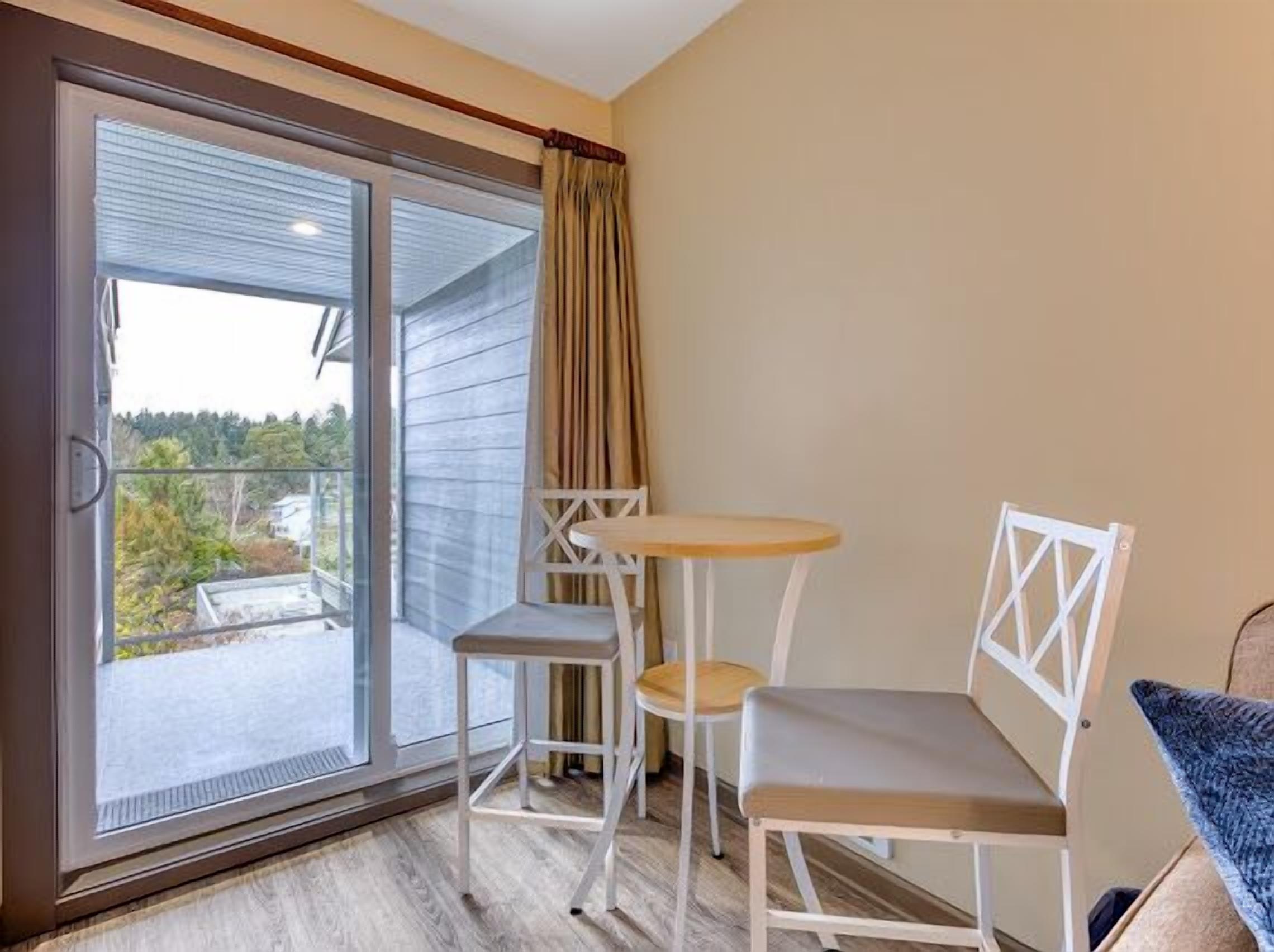 Ein Zimmer mit einer Glastür zu einem Balkon, einem kleinen runden Tisch, zwei weißen Stühlen und einer blauen, gemusterten Decke auf einem Stuhl.