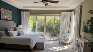 Een slaapkamer met een groot bed, een plafondventilator, een stoel, een kast en uitzicht op groen.