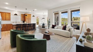 A spacious living area with a sofa, green armchairs, and a coffee table. There is a kitchen with wooden cabinets and a dining area with a table and chairs.