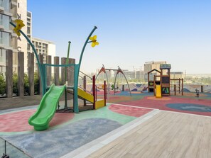 A playground with a slide, swings, and climbing structures, surrounded by buildings and trees. - The Grove - 411 Studio (Dubai)