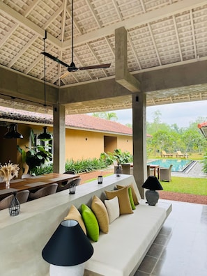 A covered outdoor seating area with a pool view, featuring a long white sofa, black lamps, and a dining table with chairs. - Raya Island Villa (Panadura)