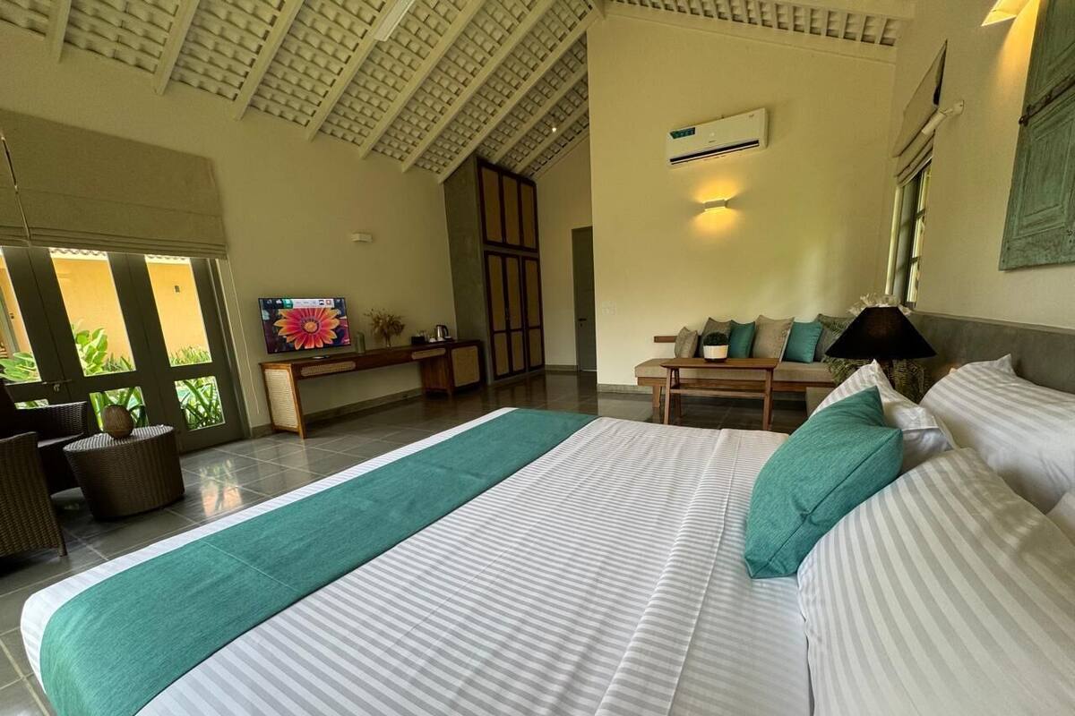 A spacious bedroom with a large bed, a view of greenery through the windows, a television, and a seating area with a table and chairs.