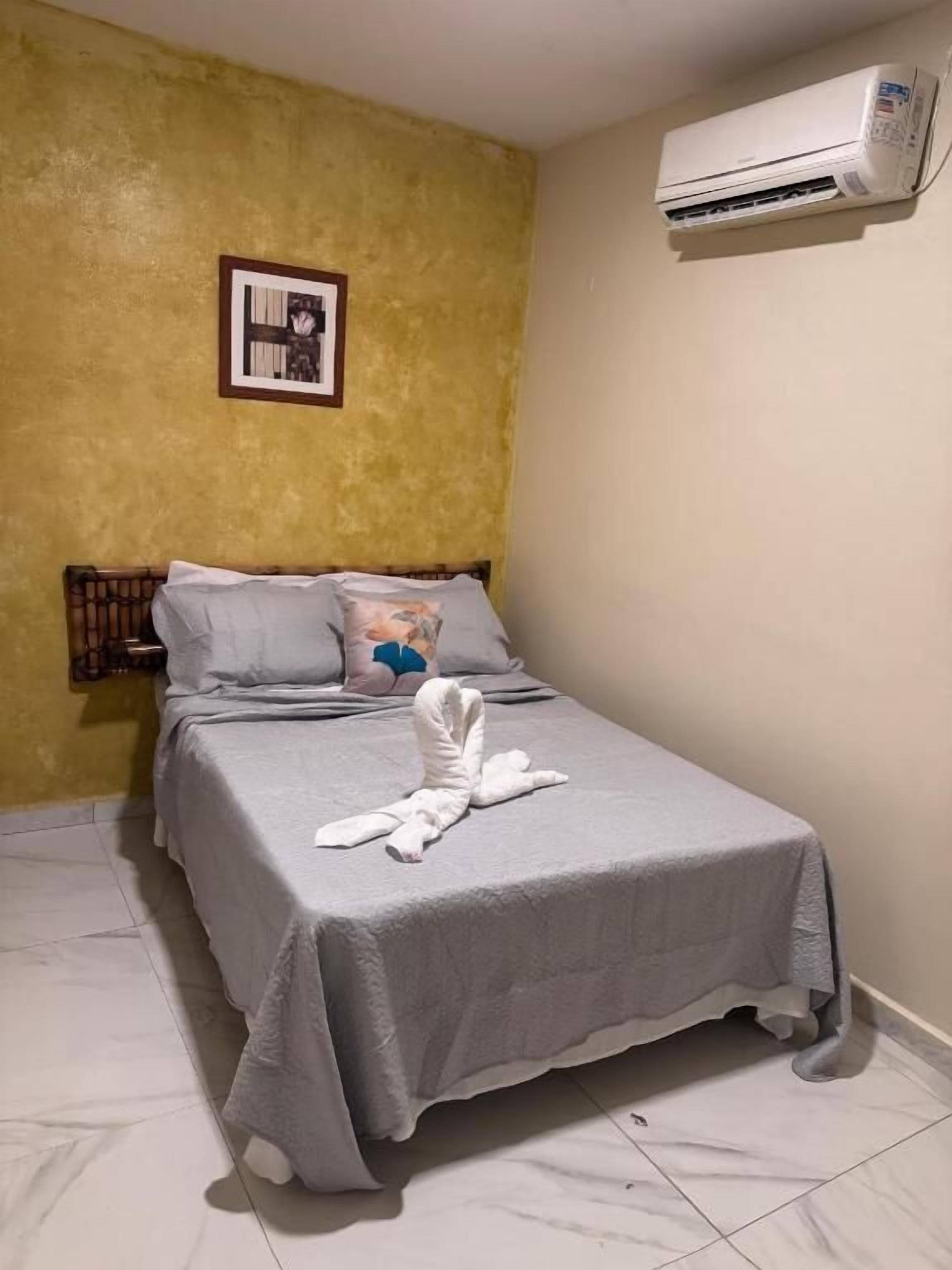 Una cama individual con una colcha gris y una toalla blanca doblada encima, un cuadro en la pared y un equipo de aire acondicionado montado sobre la cama.