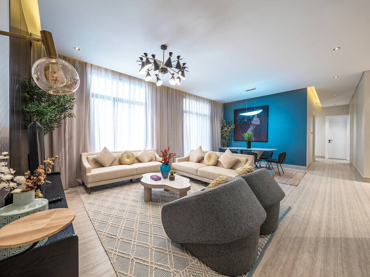 A modern living room with a beige sofa set, a round coffee table, and a grey armchair. There is a large window with sheer curtains and a blue accent wall.