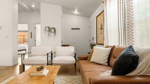 Una sala de estar con un sofá de cuero marrón, dos sillas blancas, una mesa de centro de madera y una alfombra estampada.