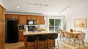 Una cocina moderna con armarios de madera, un refrigerador negro, un microondas y una cafetera. Hay una zona con taburetes de bar, con tres taburetes, y una mesa de comedor con seis sillas.