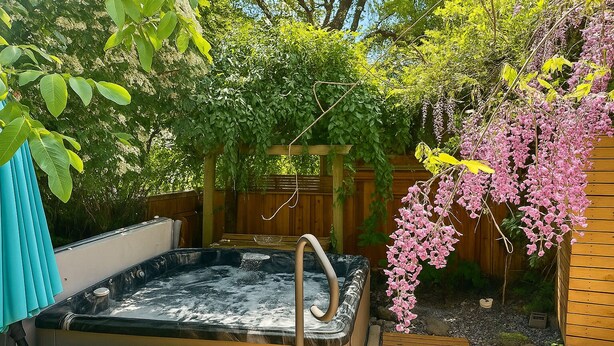 Een hottub in een tuin met houten terras, omgeven door weelderige begroeiing en bloeiende planten.