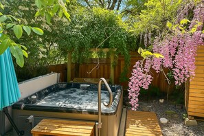 A hot tub in a garden with wooden decking, surrounded by lush greenery and flowering plants.