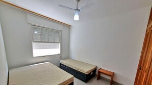 A small, sparsely furnished room with two beds, a ceiling fan, a window with blinds, and a small wooden bench. - Renovated apartment, just 250m from the sea, fast Wi-Fi, close to the Aquarium, for 4 people (Guarujá)