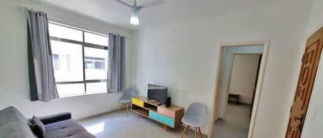 Una sala de estar con un sofá, un televisor sobre un mueble de madera, un ventilador de techo y una ventana con cortinas.