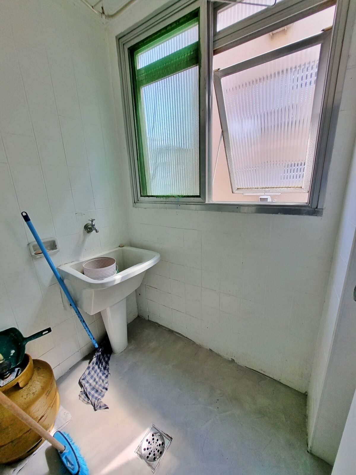 Un baño pequeño y limpio con un lavabo blanco, una ventana con persianas y utensilios de limpieza en el suelo.