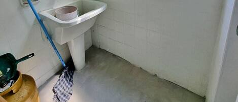Un baño pequeño y limpio con un lavabo blanco, una ventana con persianas y utensilios de limpieza en el suelo.