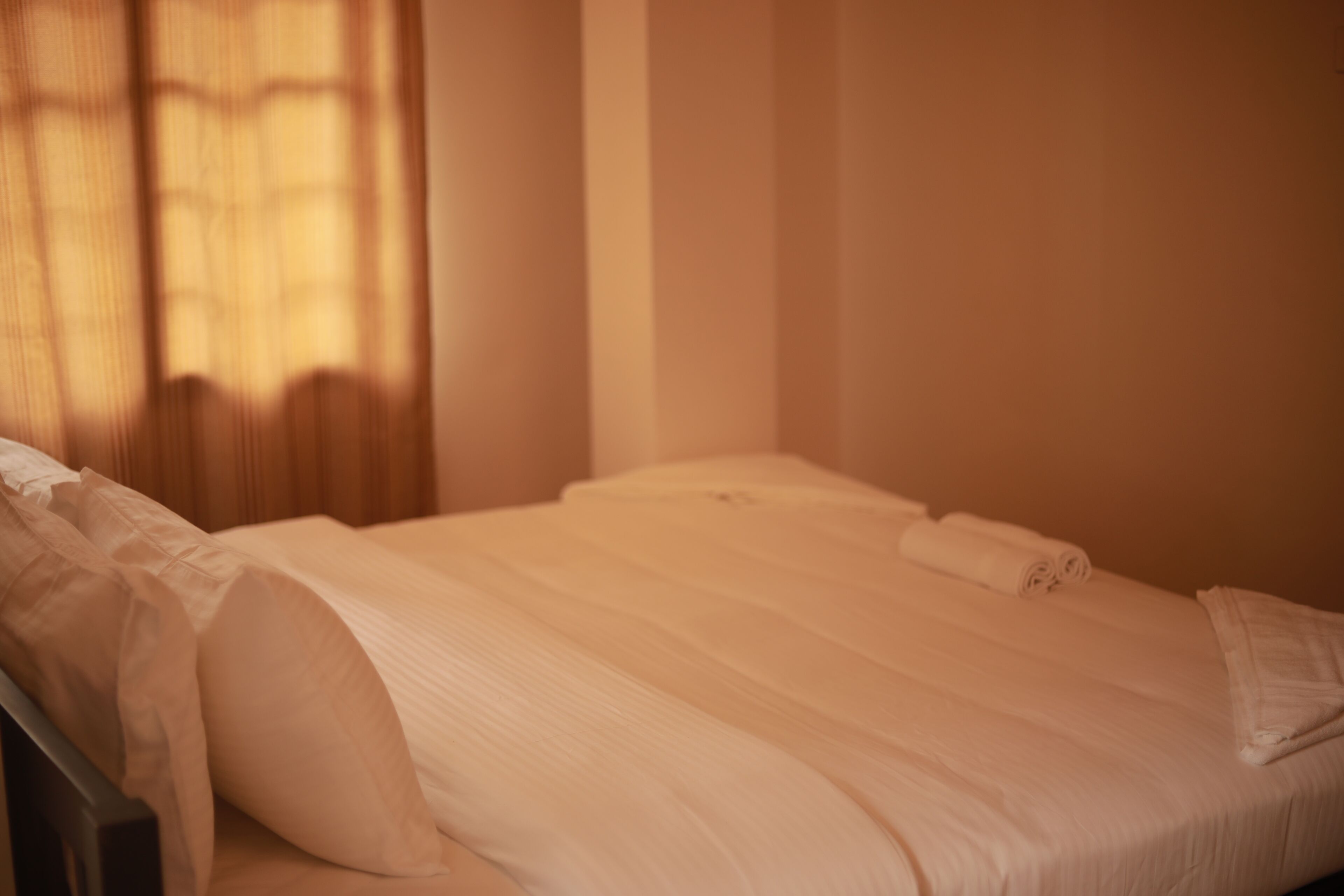 A neatly made bed with white linens in a room with a window and curtains.