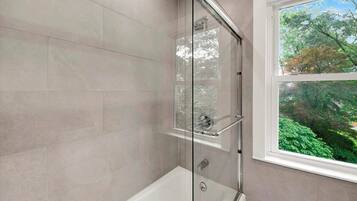 A modern bathroom with a glass-enclosed shower, a bathtub, and a window with a view of trees.