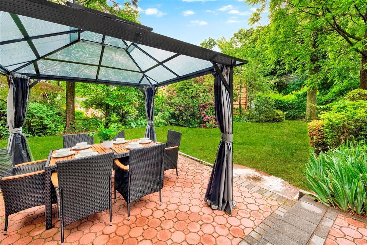 An outdoor patio with a glass-covered table and chairs, surrounded by a garden.