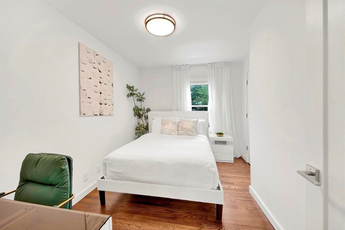A modern bedroom with a white bed, a green chair, a nightstand, and a window with curtains.